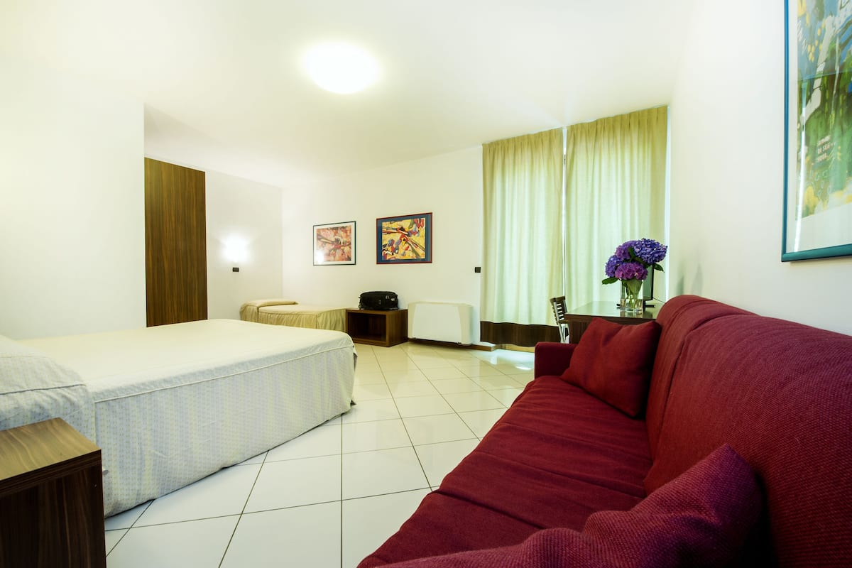 Een hotelkamer met een bed, een rode bank, een kleine tafel met een bloemenvaas, en twee ingelijste foto's aan de muur.