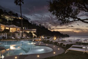Una piscina con sillas de descanso, un edificio con varios balcones y vista al océano al anochecer.