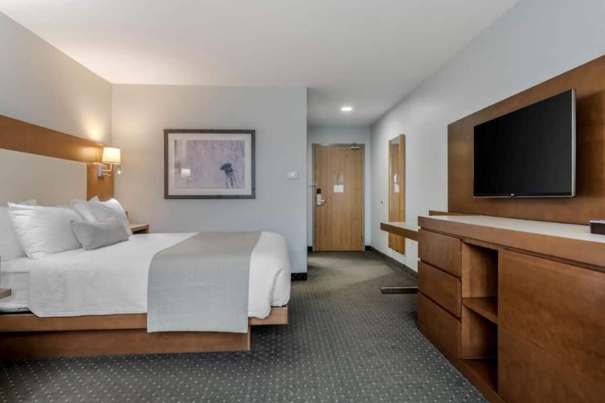 A hotel room with a bed, bedside table, lamp, artwork, TV, and a wooden dresser.