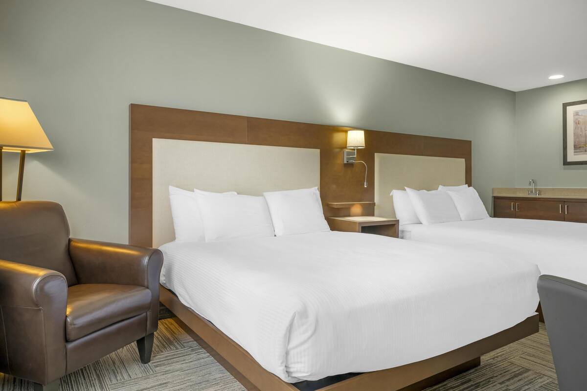 A hotel room with two beds, a brown leather chair, a nightstand with a lamp, and a wooden headboard.