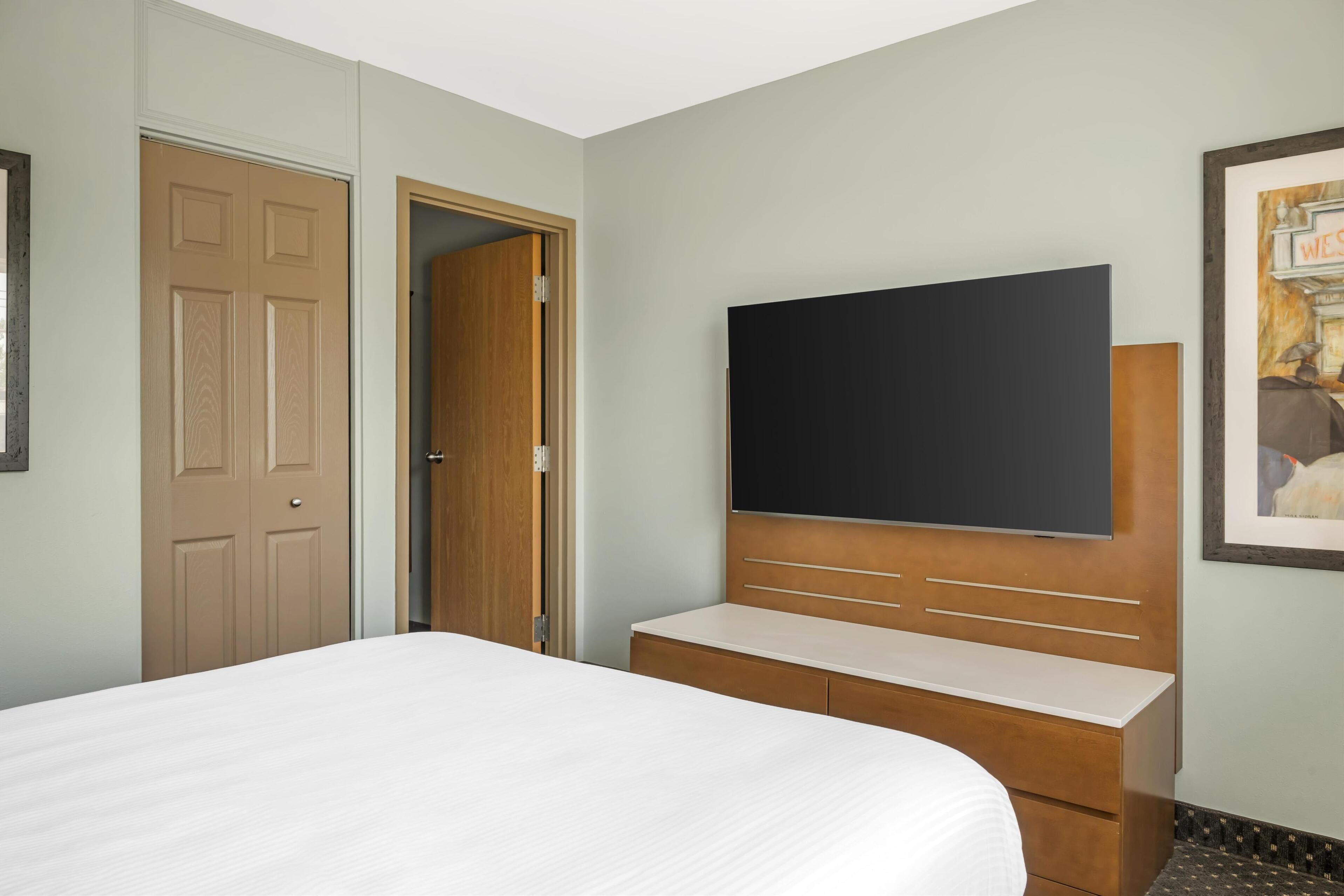 A hotel room with a bed, a wooden dresser, a flat-screen TV mounted on the wall, and a door leading to another room.