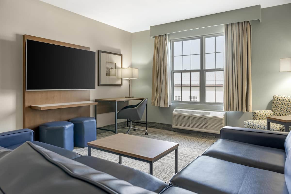 A modern hotel room with a large flat-screen TV, a desk, two blue sofas, a small table, and a window with curtains.