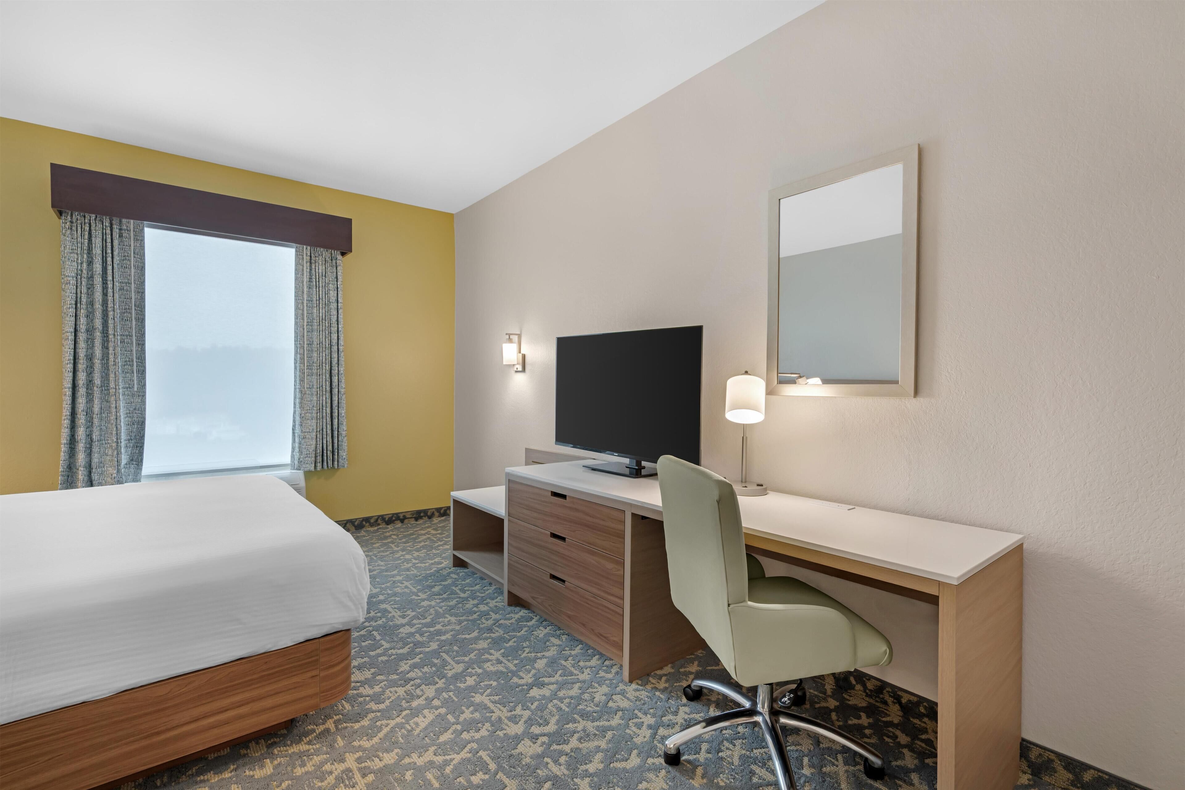 Habitación de hotel con cama, escritorio, silla, televisión y un ventanal con cortinas.