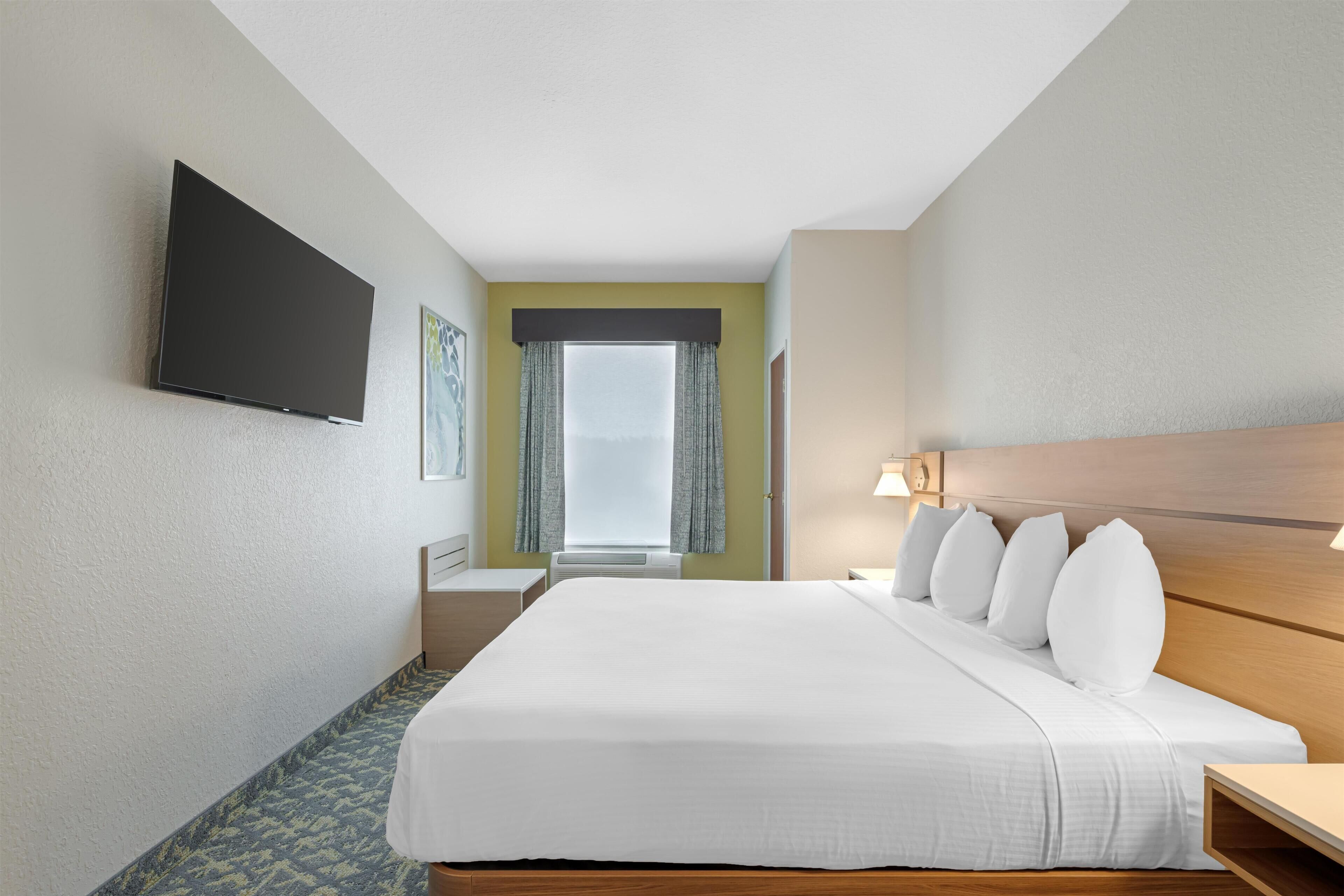 A hotel room with a large bed, a flat-screen TV mounted on the wall, a window with curtains, and a bedside table with a lamp.