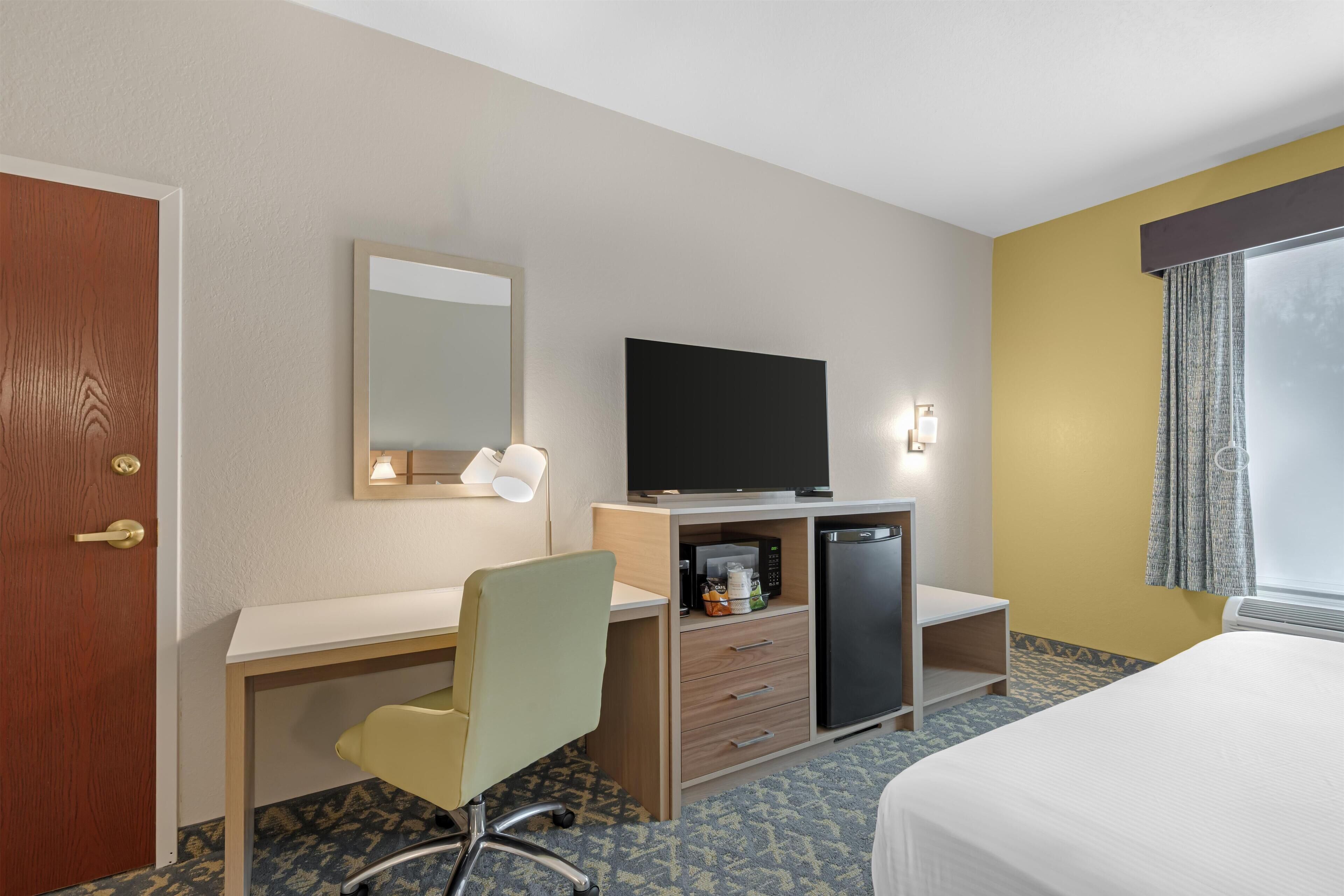 A hotel room with a wooden desk, a television, a microwave, a refrigerator, a bed, and a window with curtains.
