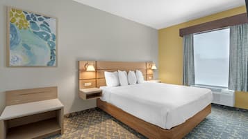 A hotel room with a large bed, bedside tables with lamps, a small desk, a large window with curtains, and a wall-mounted abstract painting.