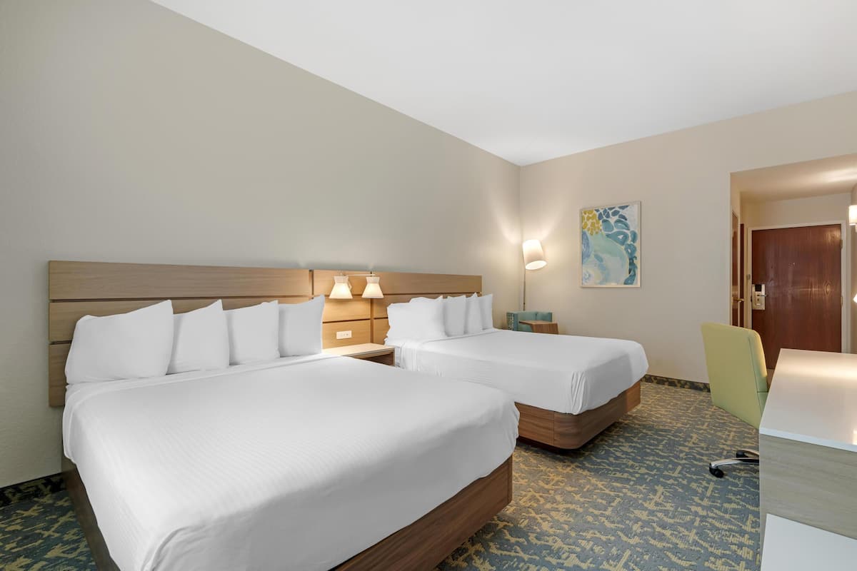 Une chambre d’hôtel avec deux lits, un bureau et une chaise. Il y a un tableau au mur et une porte à l’arrière-plan.