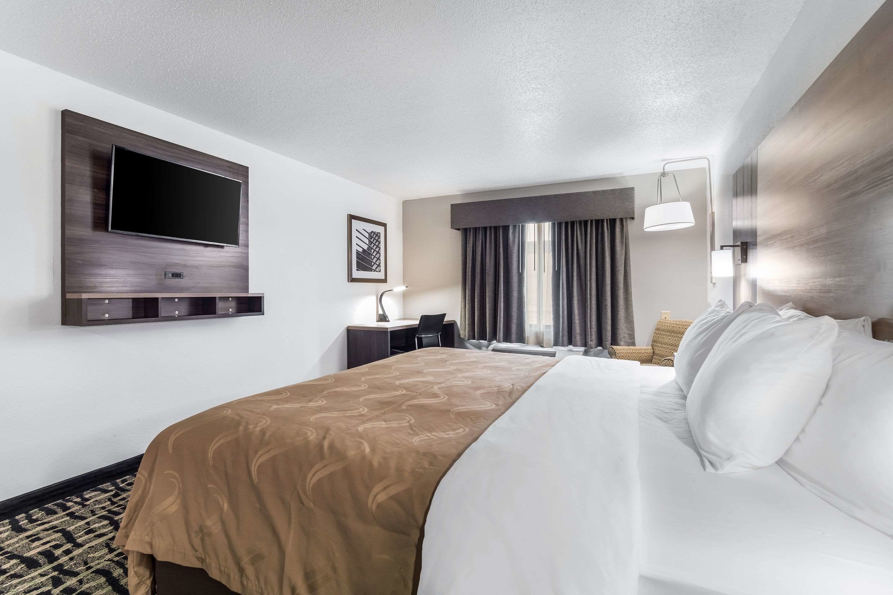 A hotel room with a large bed, a flat-screen TV mounted on the wall, a desk with a chair, and a window with curtains.