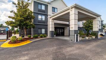 A modern hotel entrance with a covered driveway, a landscaped area with shrubs, and a clear sky.