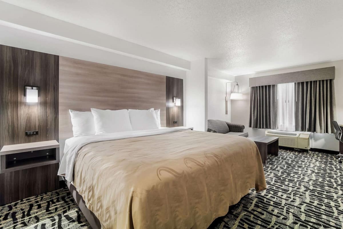 Une chambre d’hôtel avec un grand lit, des tables de chevet, un bureau, une chaise, une fenêtre avec des rideaux et un appareil de climatisation.
