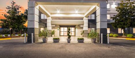 A modern hotel entrance with a covered walkway, potted plants, and a clear sky.
