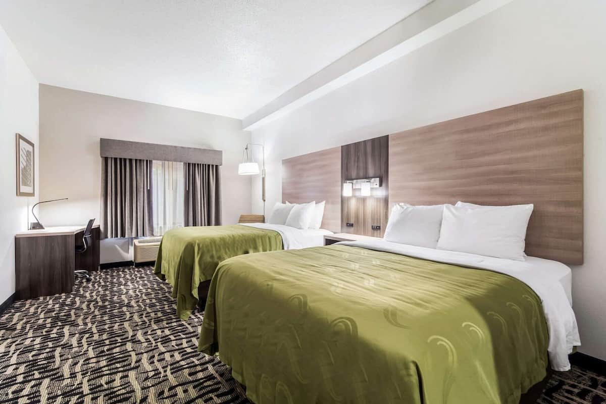 Une chambre d’hôtel avec deux lits, un bureau, une chaise, une fenêtre avec des rideaux et un tapis à motifs.