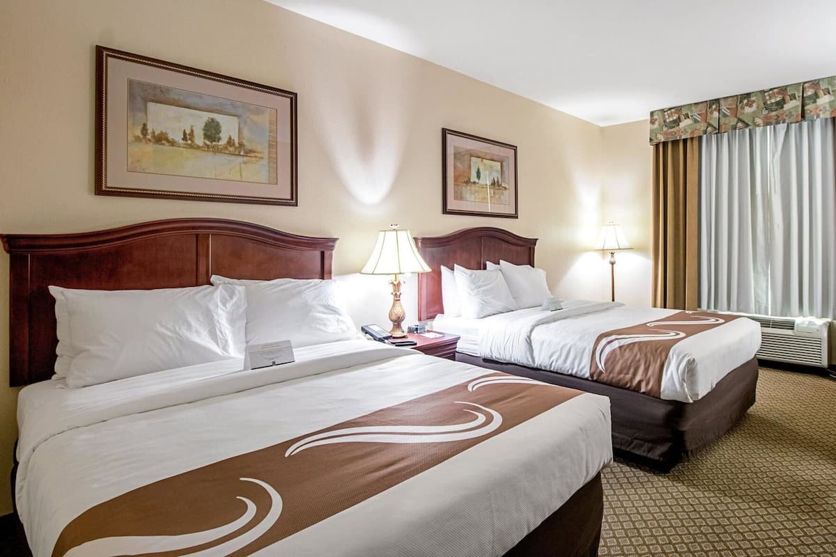A hotel room with two beds, wooden headboards, framed artwork on the walls, and a large window with curtains.