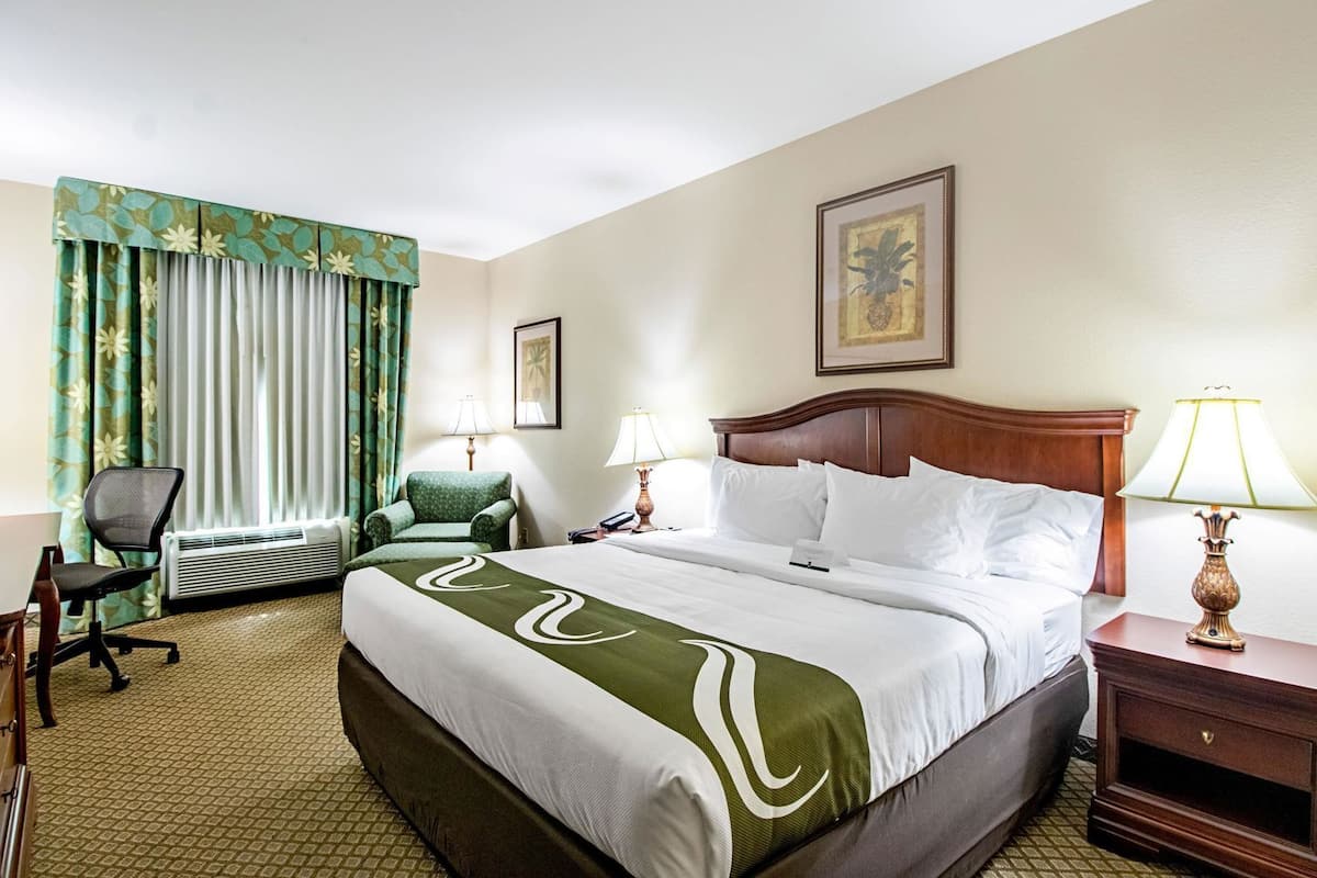 A hotel room with a large bed, two bedside tables with lamps, a chair, an air conditioning unit, and a desk with a chair.