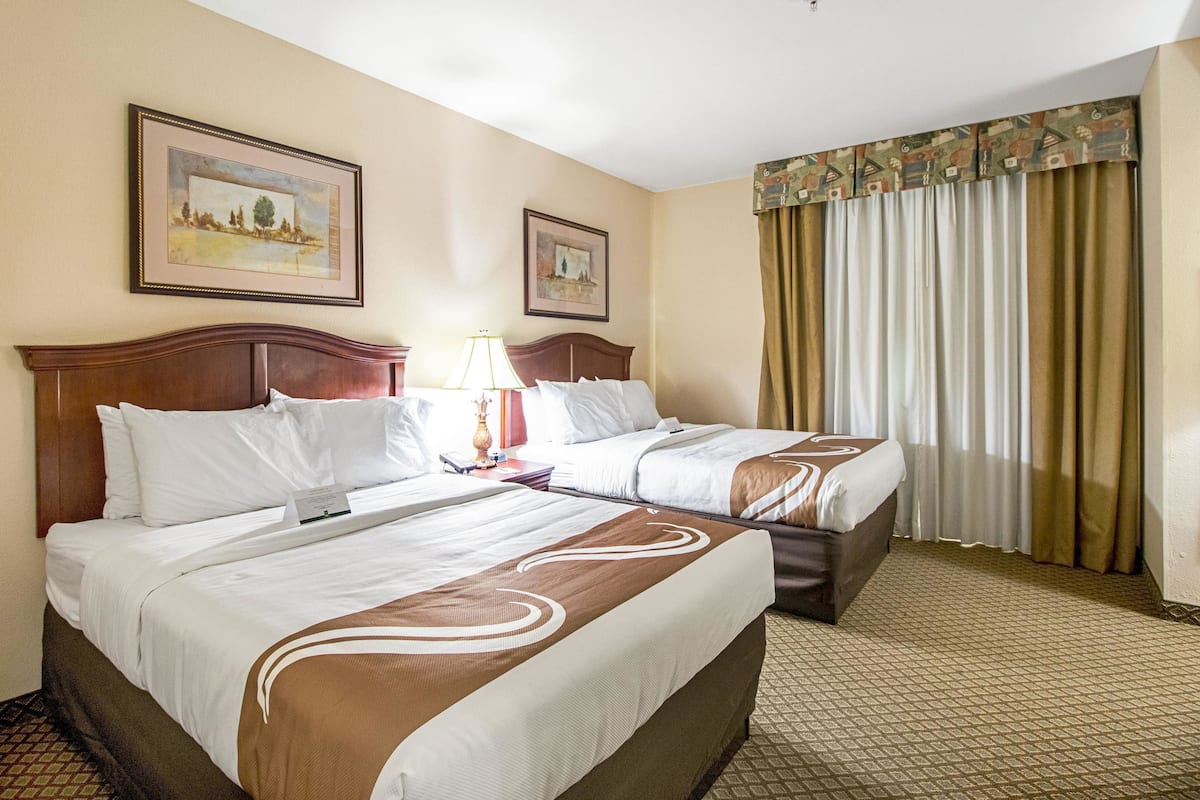 A hotel room with two beds, a nightstand, a lamp, and a window with curtains.