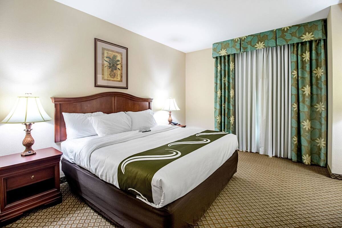 A hotel room with a bed, two bedside tables, a nightstand, a framed picture on the wall, and a window with curtains.