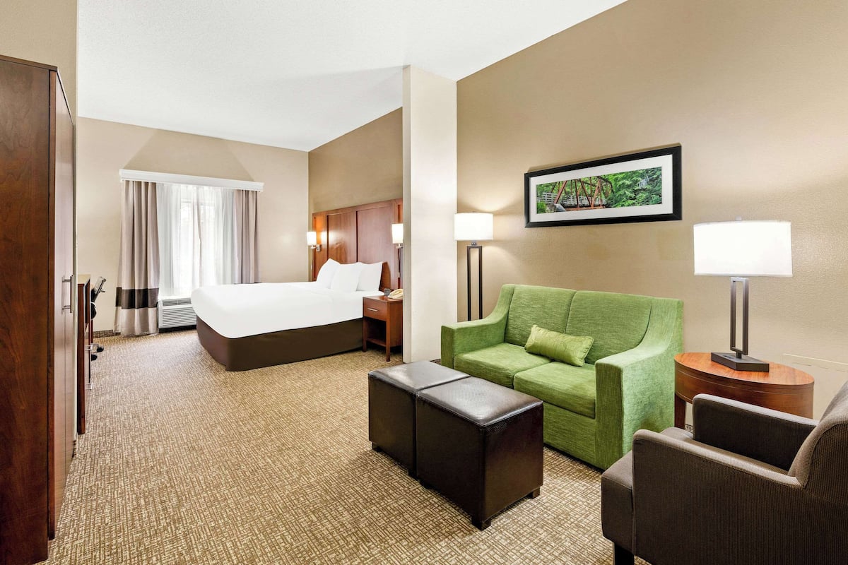 Quarto de hotel com uma cama, um sofá verde, uma poltrona marrom, uma mesinha e um abajur.