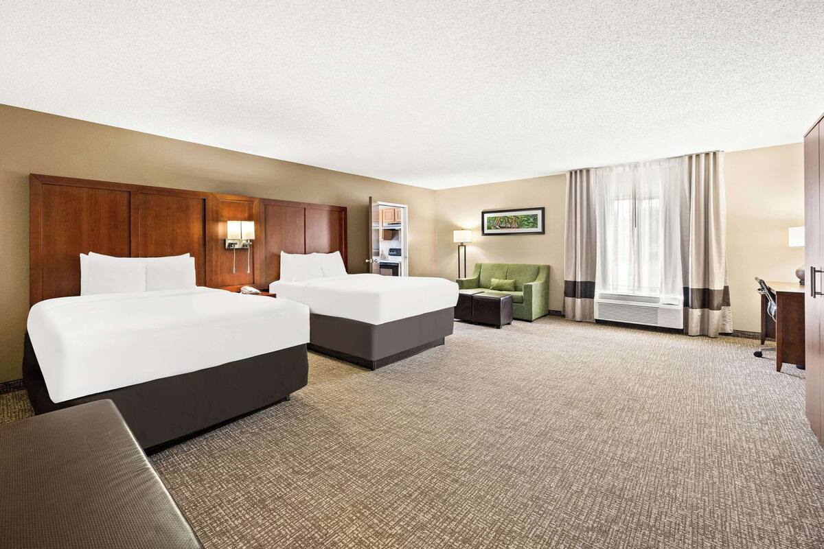 Quarto de hotel com duas camas, uma escrivaninha, uma cadeira, uma luminária e uma janela grande com cortinas.