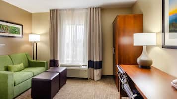 A hotel room with a green sofa, a wooden desk, a lamp, a window with curtains, and a framed picture on the wall.