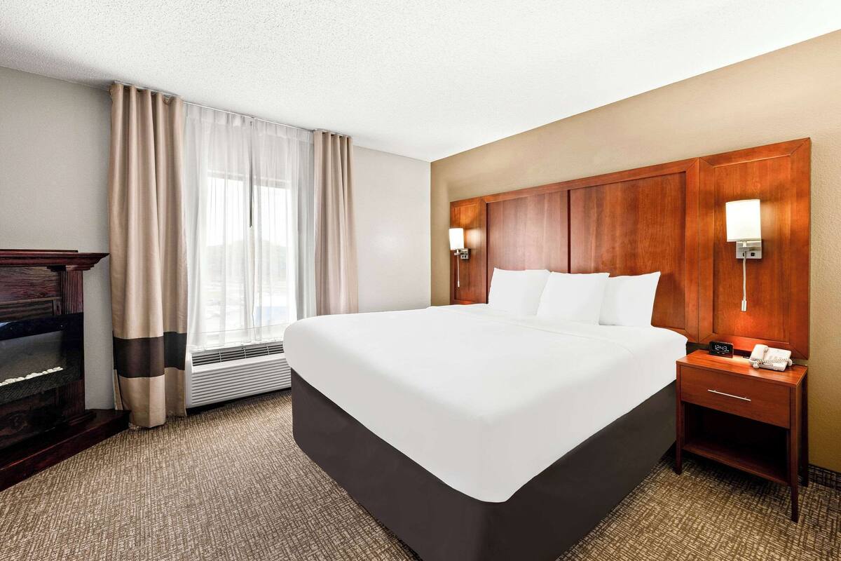 Quarto de hotel com uma cama grande, cabeceira de madeira, mesa de cabeceira com abajur e escrivaninha com telefone.