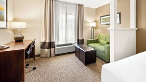 A hotel room with a desk, chair, green sofa, two ottomans, a large window with curtains, and a framed picture on the wall.