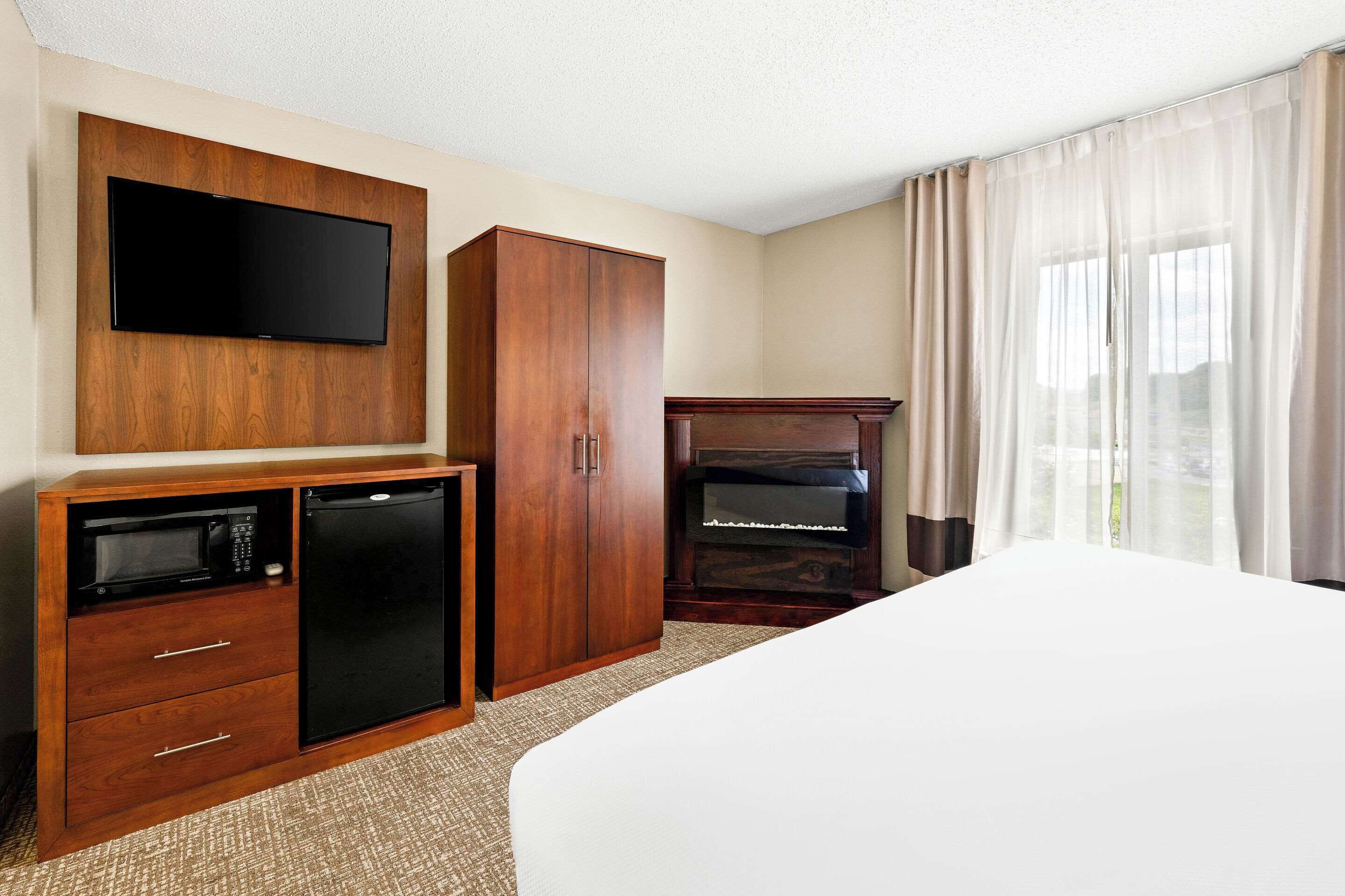 A hotel room with a bed, a TV mounted on a wooden cabinet, a minibar, a piano, and a window with curtains.
