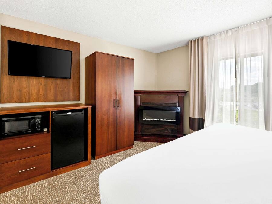 Habitación de hotel con cama, televisor montado en un mueble de madera, minibar, piano y ventana con cortinas.