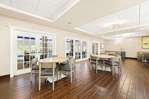 A spacious room with wooden flooring, white walls, and large windows. There are several tables and chairs arranged for dining.