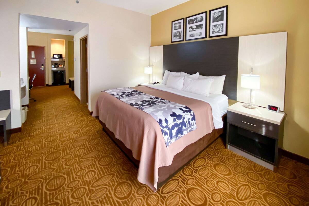 A hotel room with a large bed, a bedside table with a lamp, a small desk, and a microwave. There is a door leading to a hallway with a kitchen area.