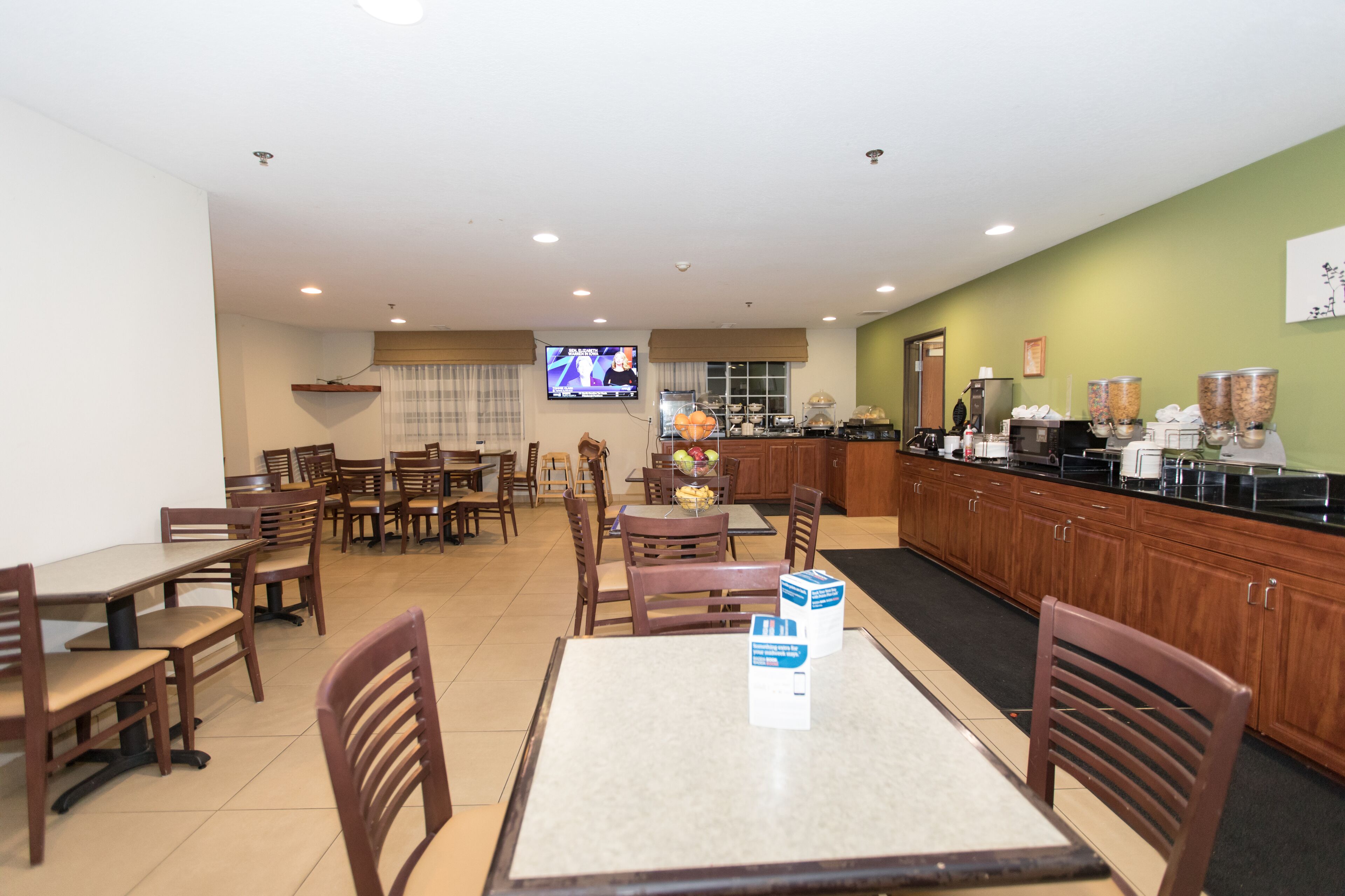 A dining area with multiple tables and chairs, a kitchen area with a coffee station, and a television displaying a news program.