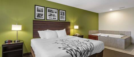 Habitación de hotel con una cama grande, una mesita de noche con lámpara, un tocador, un baño con bañera y paredes verdes.
