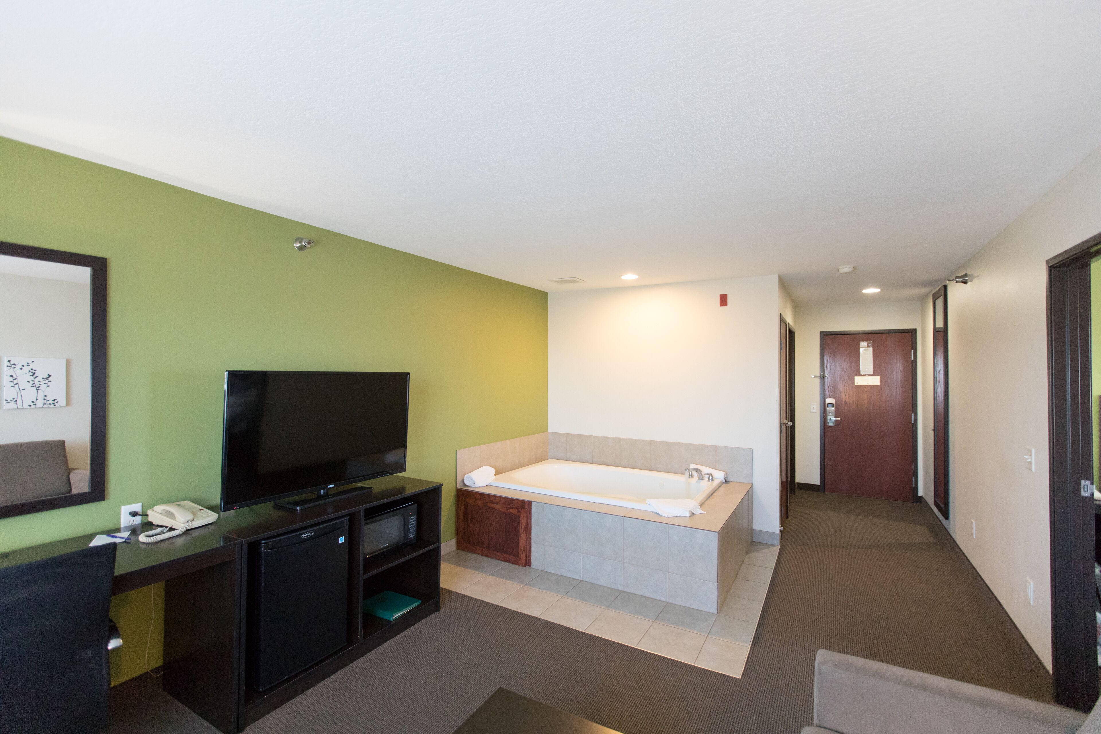 A hotel room with a flat-screen TV, a desk with a phone, a small fridge, a bathtub, and a hallway leading to another room.