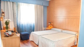 Een slaapkamer met een bed, een bureau, een stoel, een klein tafeltje met een vaas, en gordijnen.