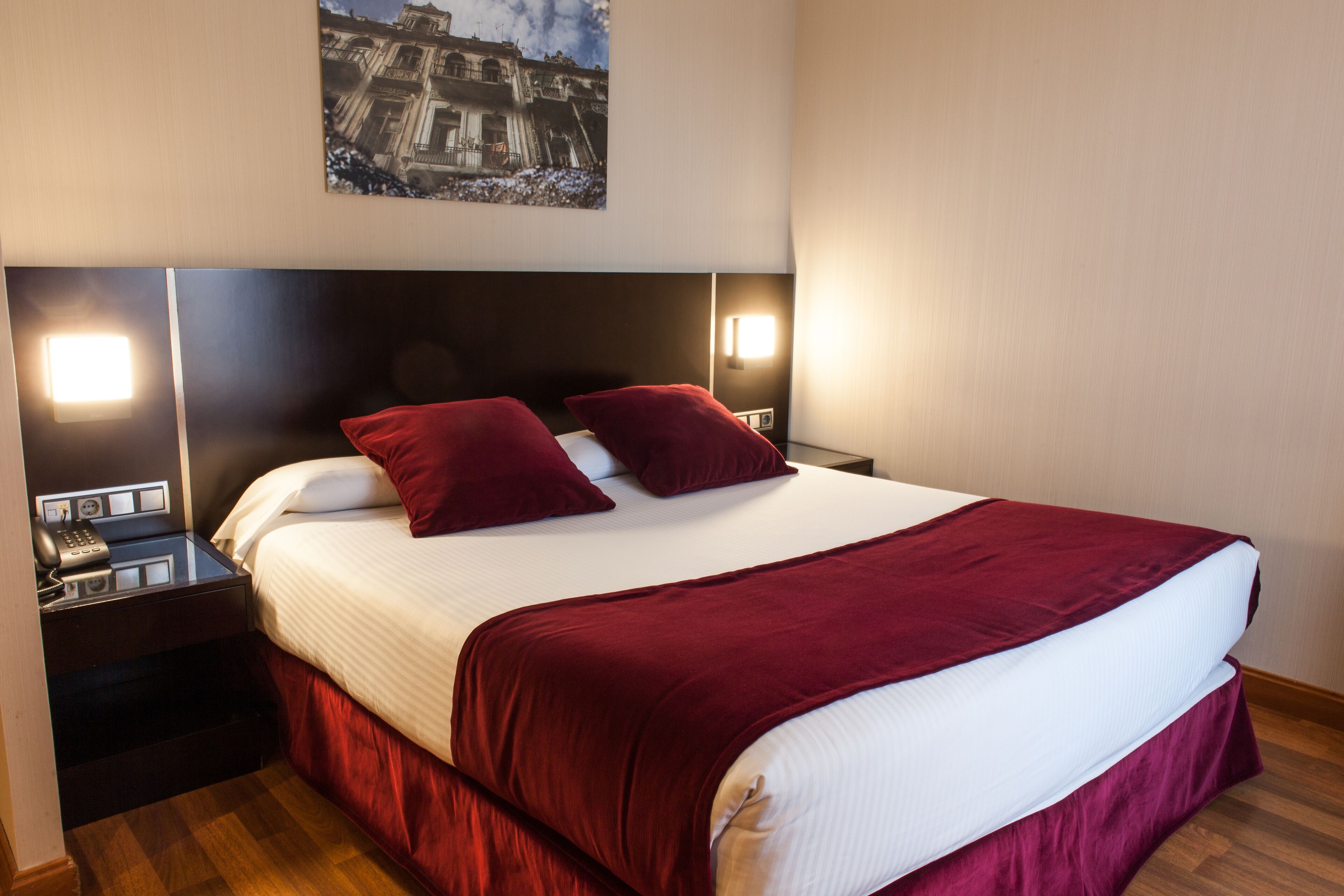 A neatly made bed with a red bedspread and pillows, a nightstand with a phone and a lamp, and a wall-mounted picture.