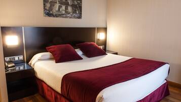 A neatly made bed with a red bedspread and pillows, a nightstand with a phone and a lamp, and a wall-mounted picture.