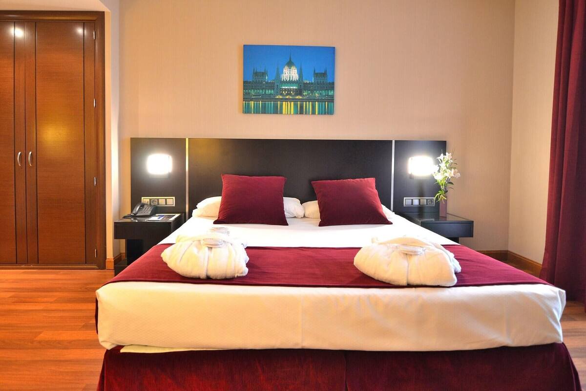 Una habitación de hotel con una cama grande, una mesita de noche con lámpara, un cuadro en la pared y un armario de madera.