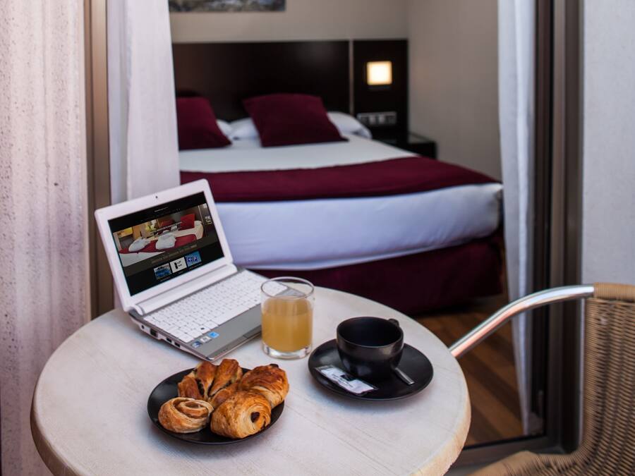 Een laptop op een klein rond tafeltje met een glas sap en croissants, een stoel, en uitzicht op een slaapkamer met een bed en een schilderij.