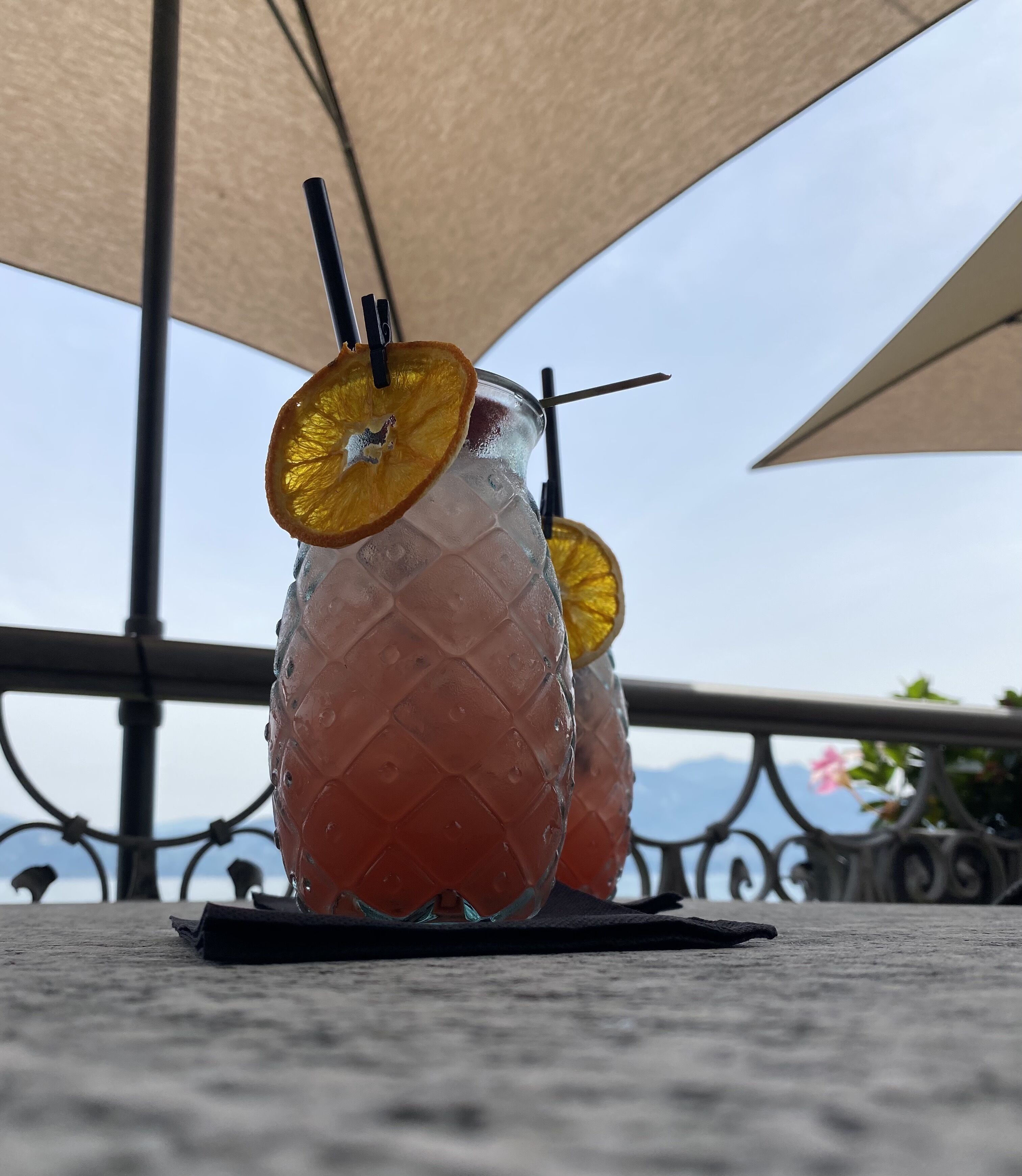 Two glasses of iced beverage with straws and orange slices on a table under a canopy.