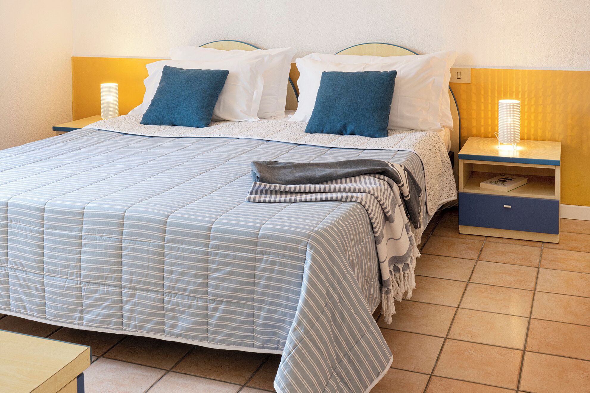 Un letto ben rifatto con copripiumino a righe, due cuscini, una coperta e un comodino con una lampada.