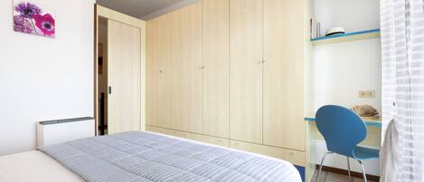 A bedroom with a bed, a wardrobe, a desk with a chair, and a shelf with books.
