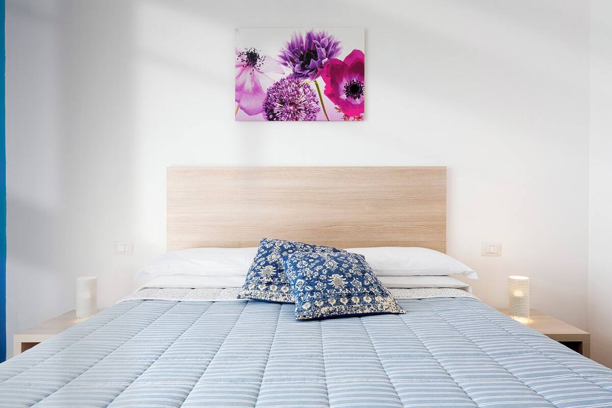 Een slaapkamer met een bed, een houten hoofdbord, twee kussens, een wandlamp en een bloemistisch kunstwerk.