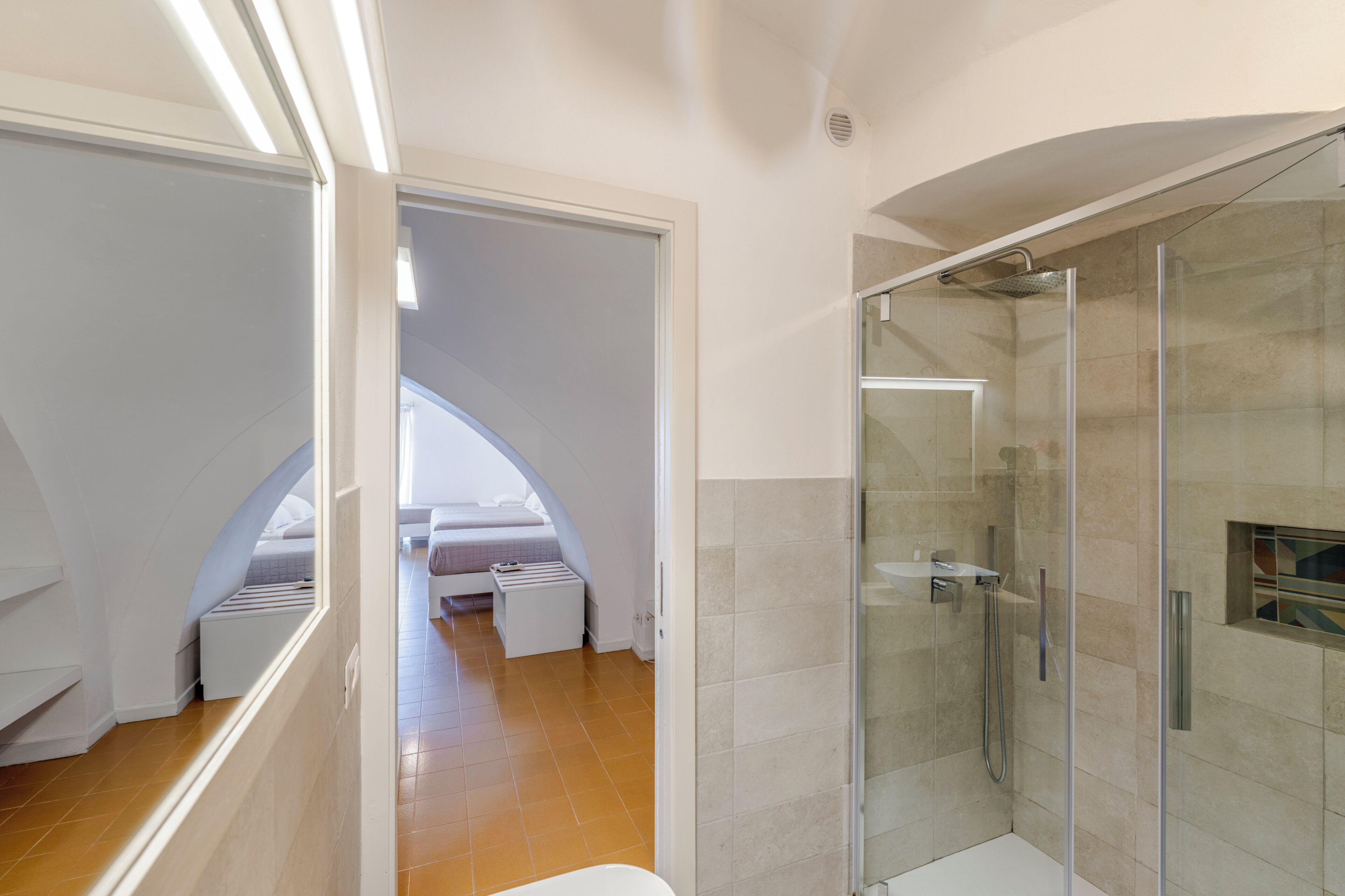 A modern bathroom with a glass shower enclosure, a white bathtub, and a visible bedroom with a curved ceiling.