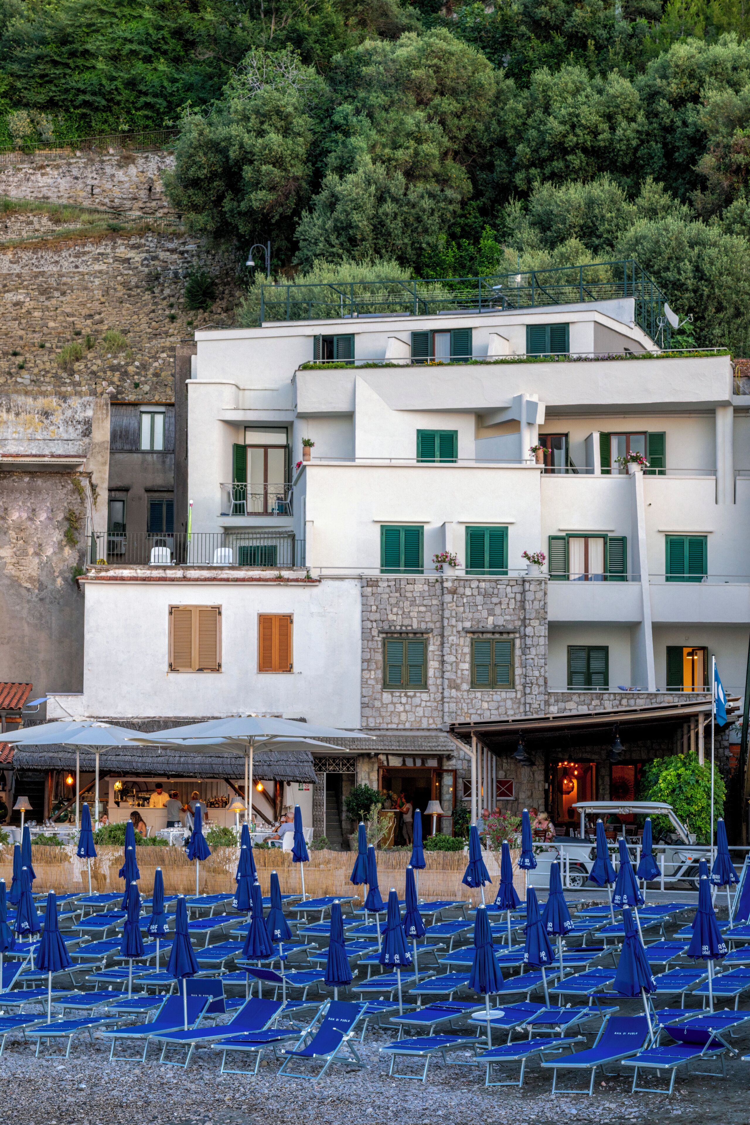 A coastal building with multiple balconies, a terrace with blue umbrellas, and a stone wall.