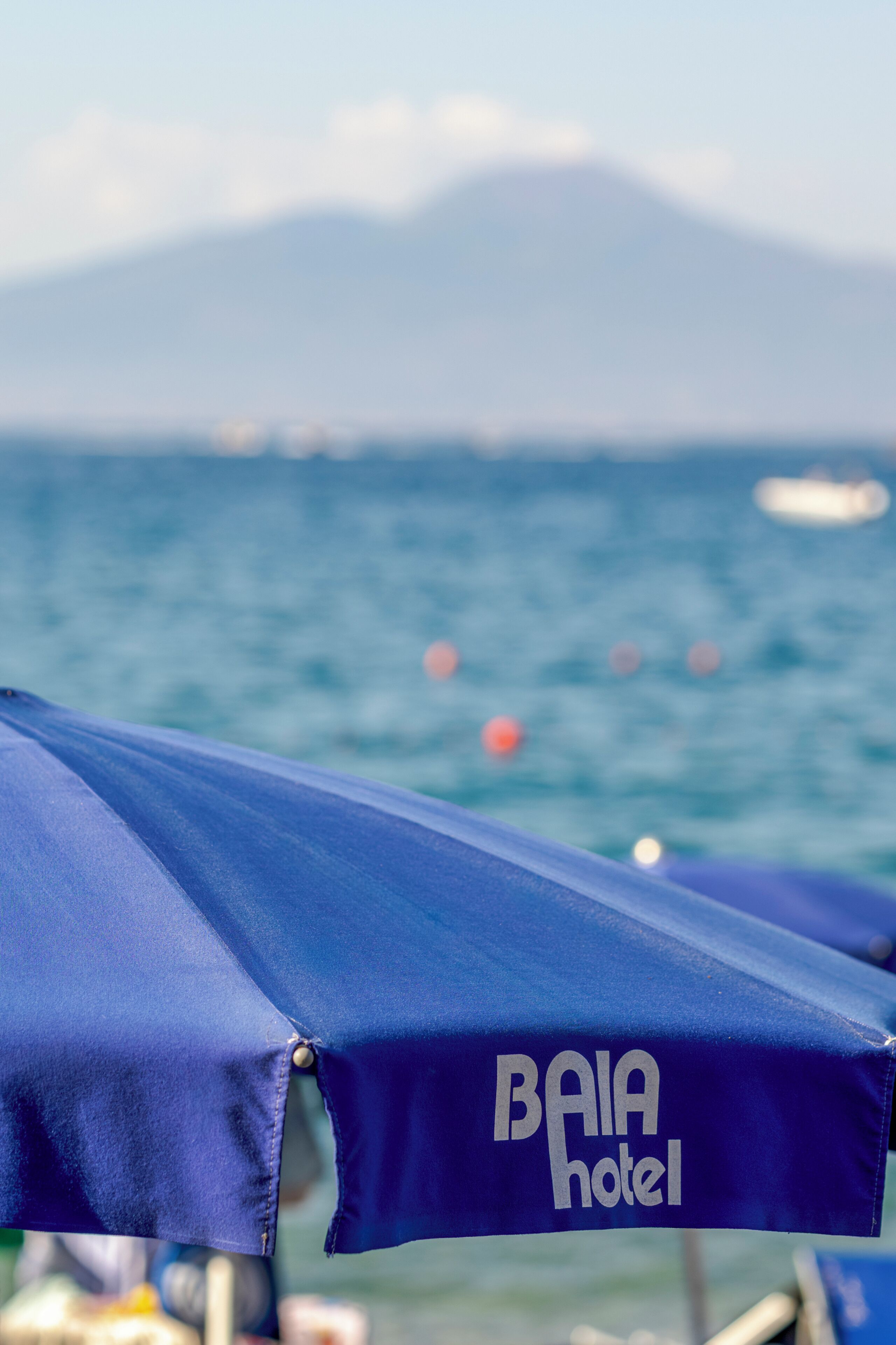 Un paraguas azul con el texto 'BAIA hotel', un cuerpo de agua y una montaña al fondo.