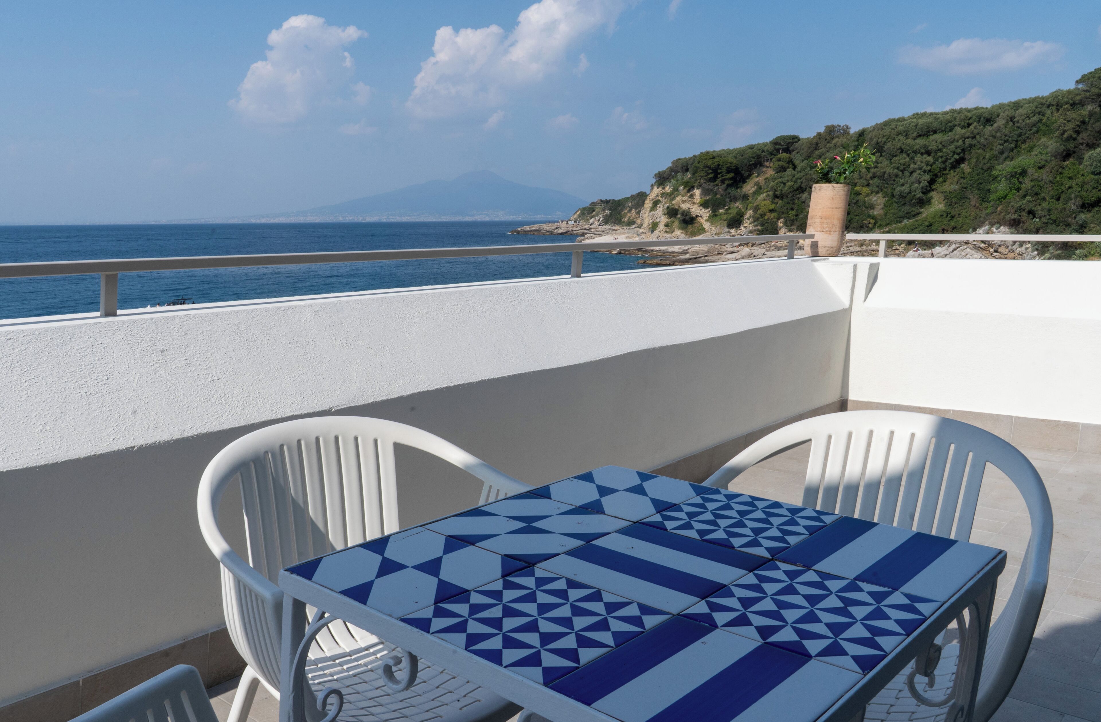 A balcony with a table and chairs, overlooking a coastal landscape with a mountain in the distance.