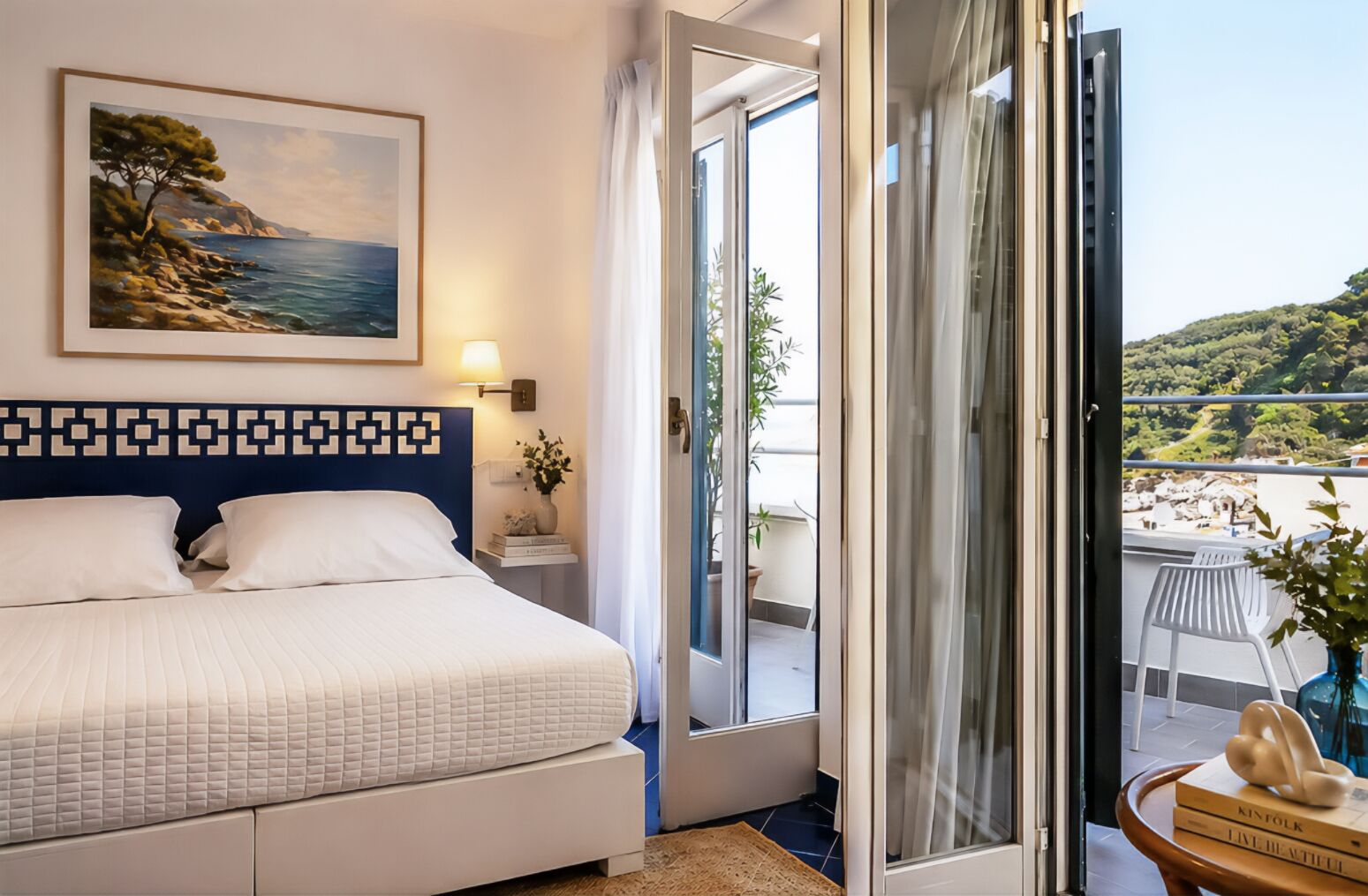 A bedroom with a bed, a nightstand, a painting, a window with a view of the sea, and a balcony with a chair.