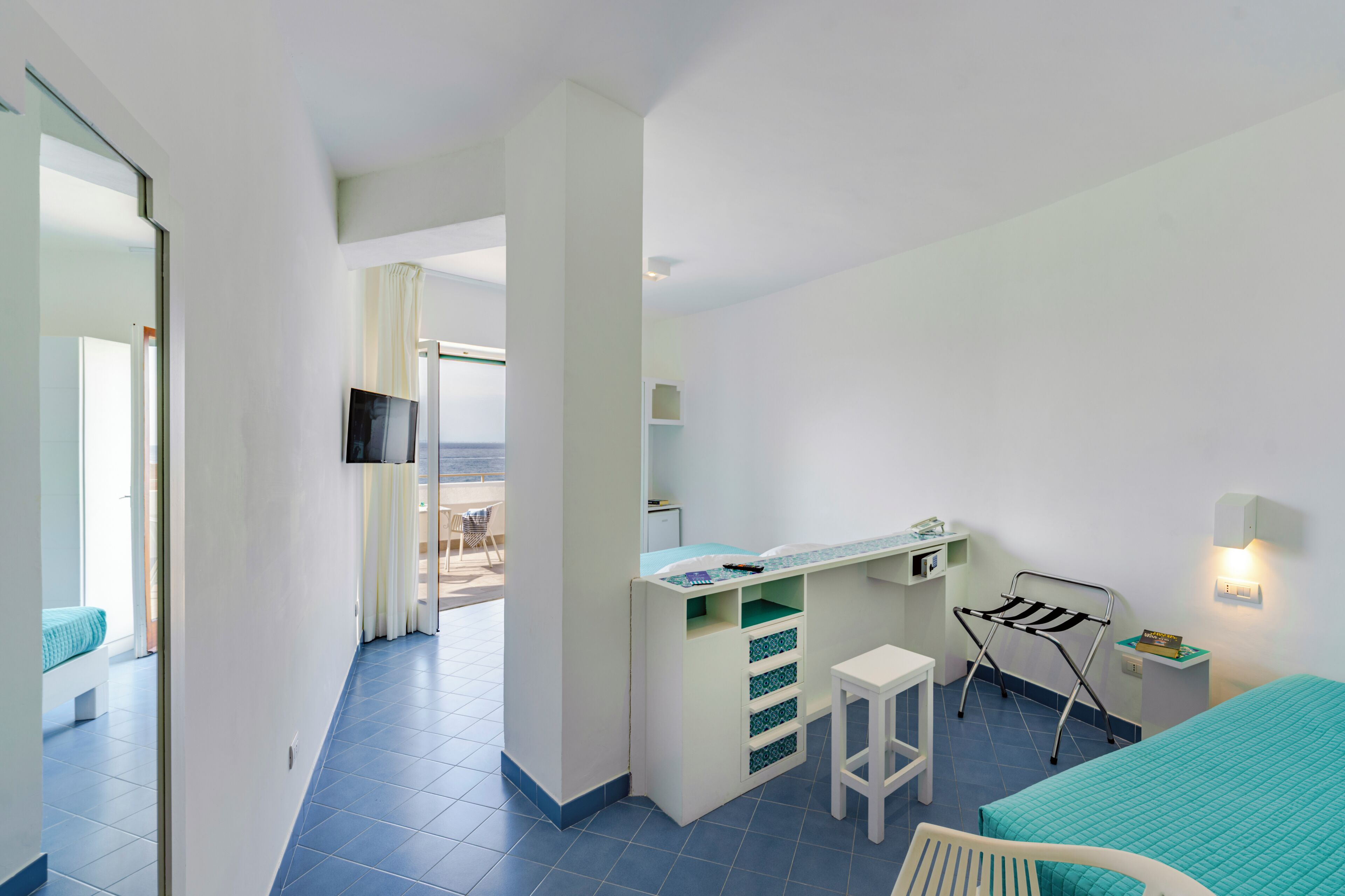 A hotel room with a bed, a desk with a chair, a TV mounted on the wall, and a view of the sea through a window.