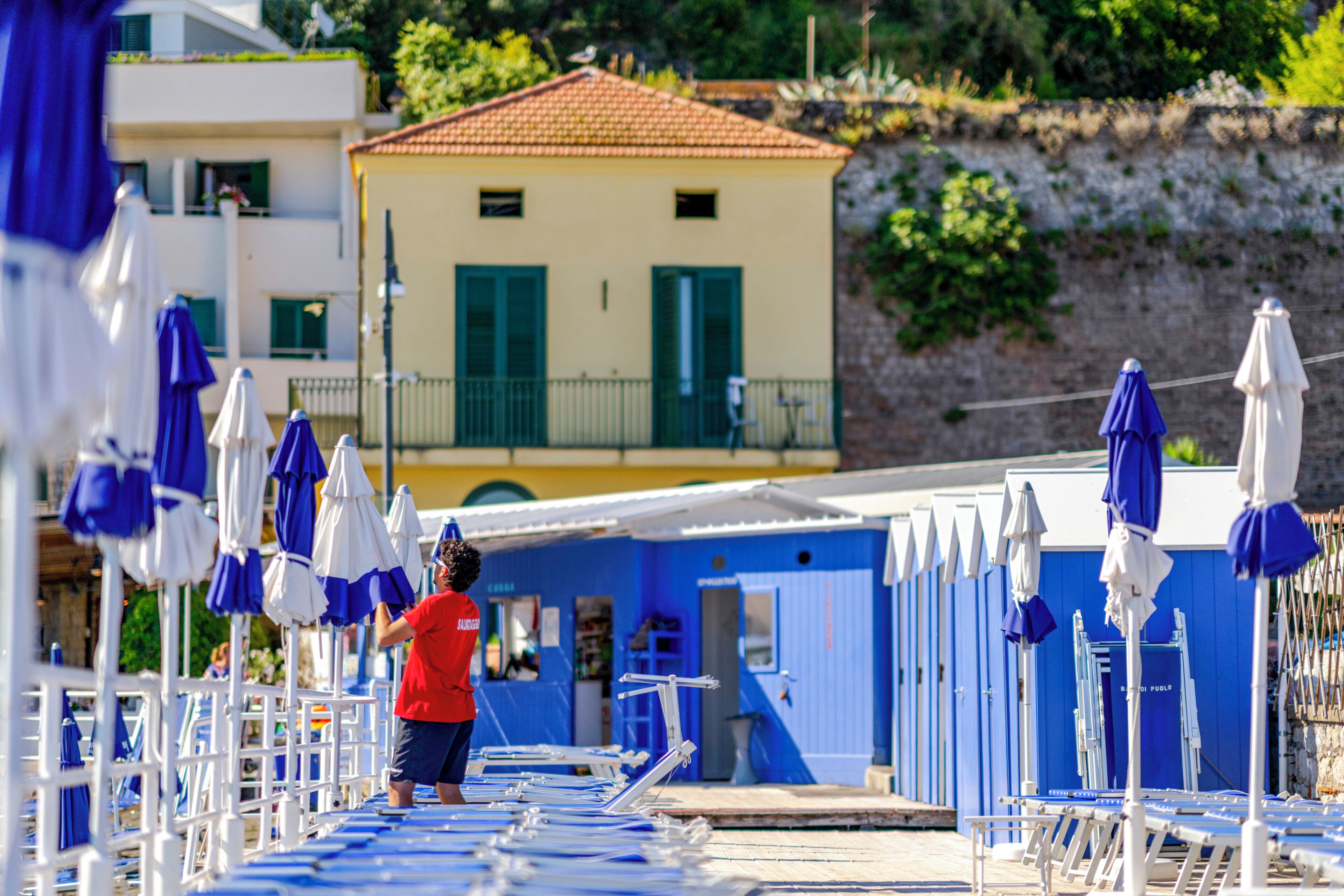 A beachside area with rows of sun loungers and umbrellas, a small building with a red roof, and a person in a red shirt.