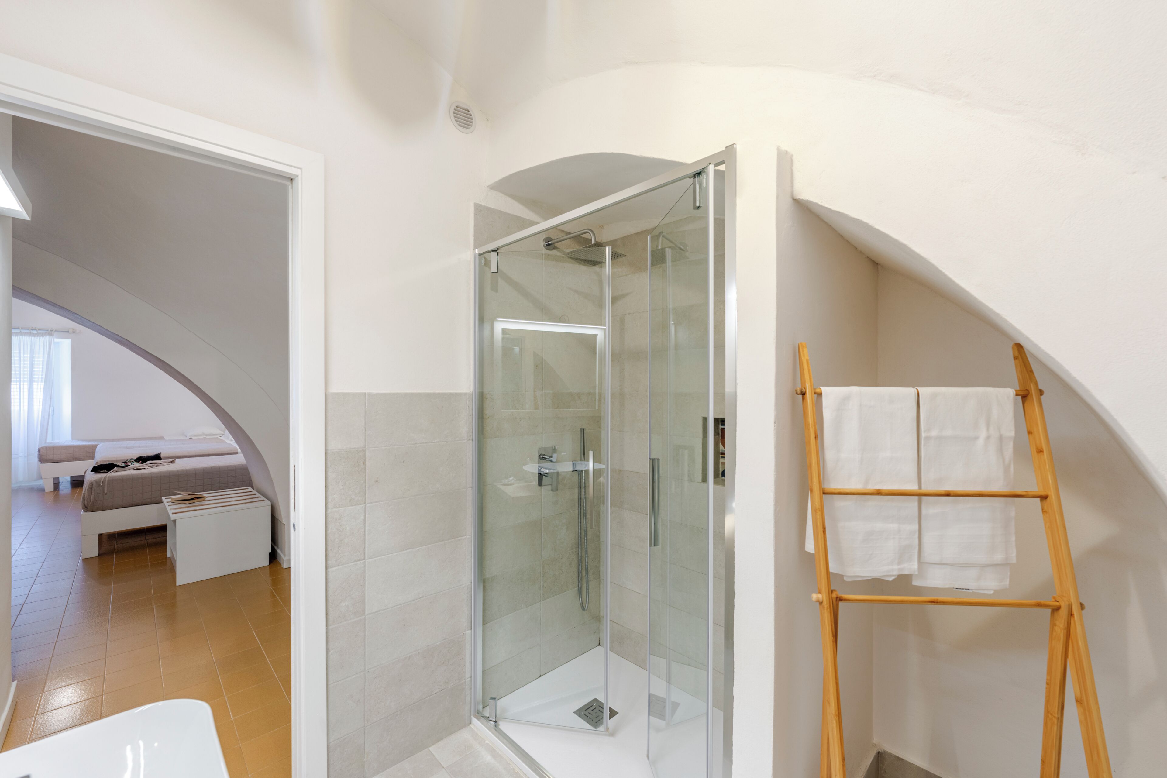 Une salle de bain moderne avec une douche à parois de verre, une échelle en bois sur laquelle sont posées des serviettes, et une chambre à coucher visible, avec un lit et une petite table.