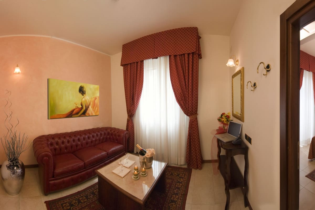 A hotel room with a red sofa, a small table with a bottle and glasses, a laptop, a bed with a red bedspread, and a mirror.